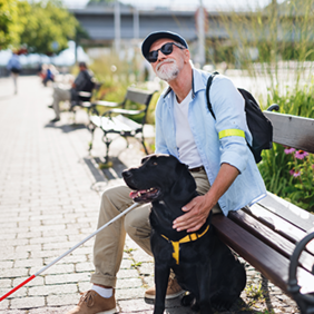 Ein Mann sitzt auf einer Parkbank aus Holz am Gehwegrand. Der Mann hat eine Sonnenbrille auf und hält einen Blindenstock in der Hand. Vor ihm sitzt ein schwarzer Hund. Der Mann guckt nach oben.