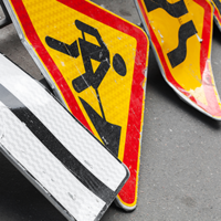 Auf dem Bild sind mehrere Verkehrsschilder zu sehen, die auf dem Boden liegen. Eines der Schilder zeigt einen schwarzen Pfeil, der nach links weist, auf weißem Hintergrund. Ein anderes Schild zeigt ein Symbol für Straßenbauarbeiten: eine stilisierte Figur, die mit einer Schaufel arbeitet, auf gelbem Hintergrund mit rotem Rand. Ein weiteres Schild zeigt eine kurvige Straße, ebenfalls auf gelbem Hintergrund mit rotem Rand.