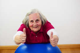 Eine ältere Frau mit grauem Haar und einem freundlichen Lächeln stützt sich auf einen blauen Gymnastikball. Sie trägt ein rotes Poloshirt über einem weißen Langarmshirt. Die Frau zeigt beide Daumen nach oben, was auf eine positive und motivierte Stimmung hinweist. Der Hintergrund ist schlicht und weiß, der Boden besteht aus hellem Holz.