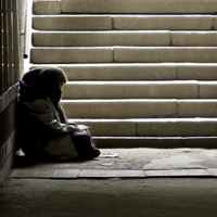 Das Bild zeigt eine Person, die in einem dunklen Durchgang oder einer Unterführung sitzt. Die Person ist in warme Kleidung gehüllt und sitzt auf dem Boden, an die Wand gelehnt. Im Hintergrund sind steinerne Treppenstufen zu sehen, die nach oben führen und von Licht durchflutet werden. 