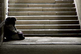 Das Bild zeigt eine Person, die in einem dunklen Durchgang oder einer Unterführung sitzt. Die Person ist in warme Kleidung gehüllt und sitzt auf dem Boden, an die Wand gelehnt. Im Hintergrund sind steinerne Treppenstufen zu sehen, die nach oben führen und von Licht durchflutet werden. 