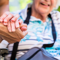 Auf dem Bild sieht man eine Nahaufnahme von zwei Händen, die sich halten. Eine Hand gehört zu einer älteren Person, die in einem Rollstuhl sitzt und ein gestreiftes Hemd trägt. Die andere Hand gehört zu einer jüngeren Person, die Pflege leistet. Im Hintergrund ist das Gesicht der älteren Person leicht unscharf zu erkennen, sie scheint zu lächeln.