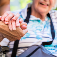 Auf dem Bild sieht man eine Nahaufnahme von zwei Händen, die sich halten. Eine Hand gehört zu einer älteren Person, die in einem Rollstuhl sitzt und ein gestreiftes Hemd trägt. Die andere Hand gehört zu einer jüngeren Person, die Pflege leistet. Im Hintergrund ist das Gesicht der älteren Person leicht unscharf zu erkennen, sie scheint zu lächeln.