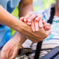 Auf dem Bild sieht man eine Nahaufnahme von zwei Händen, die sich halten. Eine Hand gehört zu einer älteren Person, die in einem Rollstuhl sitzt und ein gestreiftes Hemd trägt. Die andere Hand gehört zu einer jüngeren Person, die Pflege leistet. Im Hintergrund ist das Gesicht der älteren Person leicht unscharf zu erkennen, sie scheint zu lächeln.