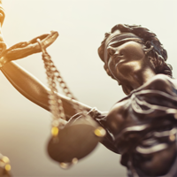 Das Bild zeigt eine Nahaufnahme einer Statue der Justitia, der römischen Göttin der Gerechtigkeit. Die Figur ist mit verbundenen Augen dargestellt, was für Unparteilichkeit steht. In ihrer rechten Hand hält sie eine Waage. Die Statue ist in einem bronzenen Farbton gehalten.