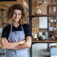 Eine Frau mit lockigem Haar steht lächelnd in einem Raum, der wie ein Atelier oder eine Werkstatt aussieht. Sie trägt ein schwarzes T-Shirt und eine graue Schürze. Im Hintergrund sind Regale mit verschiedenen Keramikstücken und Dekorationsgegenständen zu sehen. 