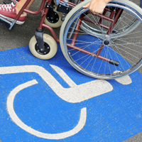 Auf dem Betonboden ist das Symbol für Rollstuhlfahrer abgebildet. Das Symbol ist in weiß auf blauen Grund. Im oberen Teil des Bildes sieht man einen Rollstuhl auf dem eine Person sitzt.