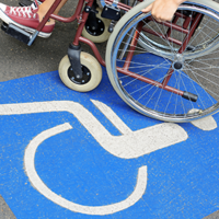 Auf dem Betonboden ist das Symbol für Rollstuhlfahrer abgebildet. Das Symbol ist in weiß auf blauen Grund. Im oberen Teil des Bildes sieht man einen Rollstuhl auf dem eine Person sitzt.