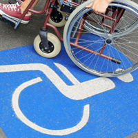 Auf dem Betonboden ist das Symbol für Rollstuhlfahrer abgebildet. Das Symbol ist in weiß auf blauen Grund. Im oberen Teil des Bildes sieht man einen Rollstuhl auf dem eine Person sitzt.