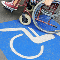 Auf dem Betonboden ist das Symbol für Rollstuhlfahrer abgebildet. Das Symbol ist in weiß auf blauen Grund. Im oberen Teil des Bildes sieht man einen Rollstuhl auf dem eine Person sitzt.