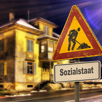Das Bild zeigt ein altes, beleuchtetes Gebäude bei Nacht. Im Vordergrund steht ein Verkehrsschild mit einem Baustellensymbol, das einen Arbeiter zeigt, der mit einer Schaufel arbeitet. Unter dem Baustellensymbol befindet sich ein weiteres Schild mit der Aufschrift "Sozialstaat". 