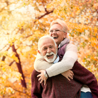 Ein älteres Paar genießt einen sonnigen Herbsttag im Freien. Die Frau, die eine Brille trägt, umarmt den Mann von hinten und beide lachen herzlich. Sie sind von herbstlich gefärbten Bäumen umgeben, deren Blätter in warmen Gelb- und Orangetönen leuchten. Der Mann trägt einen weinroten Pullover, während die Frau eine helle Jacke anhat. 