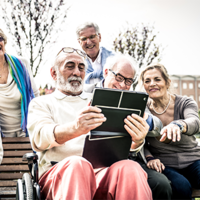Eine Gruppe älterer Menschen sitzt und steht in einem Park. Sie schauen gemeinsam auf ein Tablet, das von einem Mann in der Mitte gehalten wird. Einige der Personen lachen. Im Hintergrund sind Bäume und ein Gebäude zu sehen.