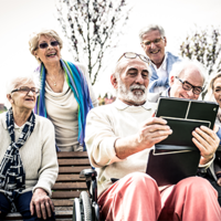 Eine Gruppe älterer Menschen sitzt und steht in einem Park. Sie schauen gemeinsam auf ein Tablet, das von einem Mann in der Mitte gehalten wird. Einige der Personen lachen. Im Hintergrund sind Bäume und ein Gebäude zu sehen.
