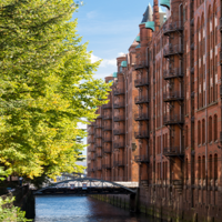 Das Foto ist von einer Brücke mit Blick auf einen Kanal in der Speicherstadt ausgenommen. Auf der linken Seite stehen hellgrüne Bäume und auf der rechten Seite sind hohe Gebäude mit Rot-Klinker-Fassade.