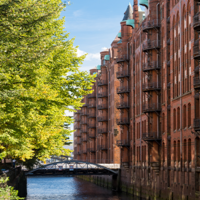 Das Foto ist von einer Brücke mit Blick auf einen Kanal in der Speicherstadt ausgenommen. Auf der linken Seite stehen hellgrüne Bäume und auf der rechten Seite sind hohe Gebäude mit Rot-Klinker-Fassade.