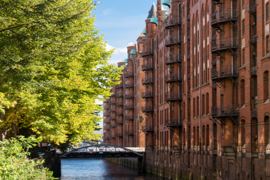 Das Foto ist von einer Brücke mit Blick auf einen Kanal in der Speicherstadt ausgenommen. Auf der linken Seite stehen hellgrüne Bäume und auf der rechten Seite sind hohe Gebäude mit Rot-Klinker-Fassade.