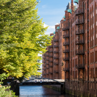 Das Foto ist von einer Brücke mit Blick auf einen Kanal in der Speicherstadt ausgenommen. Auf der linken Seite stehen hellgrüne Bäume und auf der rechten Seite sind hohe Gebäude mit Rot-Klinker-Fassade.