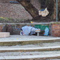Das Bild zeigt eine Szene in einem Park, in der eine Person auf einer Bank unter einer Decke schläft. Die Bank befindet sich vor einer niedrigen Backsteinmauer, hinter der ein großer Baum steht. Auf der Mauer liegt eine weitere Decke oder ein Tuch. Es gibt einige Stufen, die zu der Bank führen. Im Hintergrund sind kahle Bäume und Laub zu sehen, was auf die Herbst- oder Winterzeit hindeutet. 