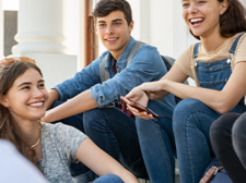 Eine Gruppe von fünf Jugendlichen sitzt auf einer Treppe und unterhält sich fröhlich. Sie lachen. Die Personen tragen lässige Kleidung, darunter Jeans, T-Shirts und Latzhosen. Im Hintergrund sind Säulen und ein Gebäude zu sehen.