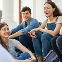 Eine Gruppe von fünf Jugendlichen sitzt auf einer Treppe und unterhält sich fröhlich. Sie lachen. Die Personen tragen lässige Kleidung, darunter Jeans, T-Shirts und Latzhosen. Im Hintergrund sind Säulen und ein Gebäude zu sehen.