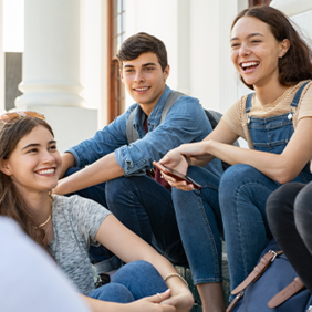 Eine Gruppe von fünf Jugendlichen sitzt auf einer Treppe und unterhält sich fröhlich. Sie lachen. Die Personen tragen lässige Kleidung, darunter Jeans, T-Shirts und Latzhosen. Im Hintergrund sind Säulen und ein Gebäude zu sehen.