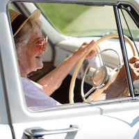 Eine ältere Frau sitzt lächelnd am Steuer eines weißen Oldtimers. Sie trägt einen Strohhut und eine Sonnenbrille. Die Sonne scheint durch das Autofenster und beleuchtet ihr Gesicht. Im Hintergrund ist unscharf eine grüne Wiese zu erkennen. Die Frau wirkt entspannt und genießt offensichtlich die Fahrt in dem klassischen Fahrzeug.