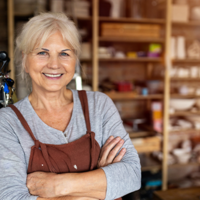 Ältere Frau mit grauen Haaren steht lächelnd in einer Werkstatt oder einem Atelier. Sie trägt ein graues Oberteil und eine braune Schürze und hat die Arme vor der Brust verschränkt. Im Hintergrund sind Regale mit Materialien und Werkzeugen zu sehen.
