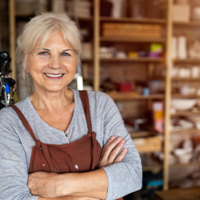 Ältere Frau mit grauen Haaren steht lächelnd in einer Werkstatt oder einem Atelier. Sie trägt ein graues Oberteil und eine braune Schürze und hat die Arme vor der Brust verschränkt. Im Hintergrund sind Regale mit Materialien und Werkzeugen zu sehen.