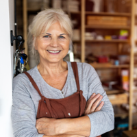 Ältere Frau mit grauen Haaren steht lächelnd in einer Werkstatt oder einem Atelier. Sie trägt ein graues Oberteil und eine braune Schürze und hat die Arme vor der Brust verschränkt. Im Hintergrund sind Regale mit Materialien und Werkzeugen zu sehen.