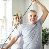 Ein älterer Mann in einem grauen T-Shirt führt eine Dehnübung mit einem blauen Widerstandsband durch. Eine Frau, möglicherweise eine Physiotherapeutin, steht hinter ihm und unterstützt ihn bei der Übung. Im Hintergrund ist ein helles Zimmer mit einem großen Fenster zu sehen, durch das Tageslicht hereinfällt. 