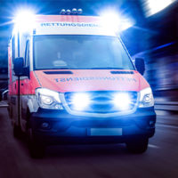 Ein Rettungswagen fährt mit eingeschaltetem Blaulicht und Sirene. Das Fahrzeug ist in den Farben Rot und Weiß gehalten und trägt die Aufschrift Rettungsdienst auf der Front. Die Scheinwerfer und das Blaulicht leuchten hell. 