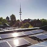 Im Vordergrund sind Solaranlagen auf einem Dach zu sehen. Im Hintergrund Bäume und in der Mitte ein Fernsehturm.