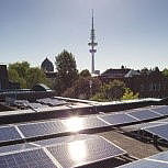 Im Vordergrund sind Solaranlagen auf einem Dach zu sehen. Im Hintergrund Bäume und in der Mitte ein Fernsehturm.