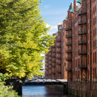 Das Foto ist von einer Brücke mit Blick auf einen Kanal in der Speicherstadt ausgenommen. Auf der linken Seite stehen hellgrüne Bäume und auf der rechten Seite sind hohe Gebäude mit Rot-Klinker-Fassade.