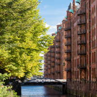Das Foto ist von einer Brücke mit Blick auf einen Kanal in der Speicherstadt ausgenommen. Auf der linken Seite stehen hellgrüne Bäume und auf der rechten Seite sind hohe Gebäude mit Rot-Klinker-Fassade.
