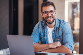 Ein Mann mit Brille und Bart sitzt an einem Schreibtisch. Er trägt ein hellblaues Jeanshemd über einem weißen T-Shirt und lächelt in die Kamera. Vor ihm steht ein aufgeklappter Laptop, und auf dem Tisch liegen verschiedene Gegenstände, darunter ein Notizbuch und ein Stift. 