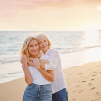 Das Bild zeigt zwei Frauen, die sich an einem Strand umarmen. Die jüngere Frau steht vorne und trägt ein weißes T-Shirt und eine blaue Jeans. Die ältere Frau steht hinter ihr und hat ihre Arme um die Schultern der jüngeren Frau gelegt. Beide lächeln glücklich in die Kamera. Im Hintergrund ist das Meer zu sehen.