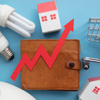 Das Bild zeigt eine braune Geldbörse, auf der ein roter Pfeil in Form eines steigenden Diagramms platziert ist. Um die Geldbörse herum sind verschiedene Gegenstände angeordnet: zwei Glühbirnen, ein Miniatur-Einkaufswagen und zwei kleine Häuser. Der Hintergrund ist hellblau. Das Bild symbolisiert steigende Kosten oder Preise in verschiedenen Lebensbereichen wie Energie, Wohnen und Konsumgüter.