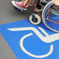 Auf dem Betonboden ist das Symbol für Rollstuhlfahrer abgebildet. Das Symbol ist in weiß auf blauen Grund. Im oberen Teil des Bildes sieht man einen Rollstuhl auf dem eine Person sitzt.