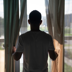 Ein Mann steht in einem hellen Raum vor einem großen Fenster und hält die Vorhänge zur Seite. Er trägt ein weißes T-Shirt und blickt nach draußen, wo man unscharf eine grüne Landschaft und Gebäude im Hintergrund erkennen kann. Das Licht von draußen erhellt den Raum leicht, während die Vorhänge teilweise geöffnet sind.