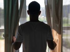 Ein Mann steht in einem hellen Raum vor einem großen Fenster und hält die Vorhänge zur Seite. Er trägt ein weißes T-Shirt und blickt nach draußen, wo man unscharf eine grüne Landschaft und Gebäude im Hintergrund erkennen kann. Das Licht von draußen erhellt den Raum leicht, während die Vorhänge teilweise geöffnet sind.