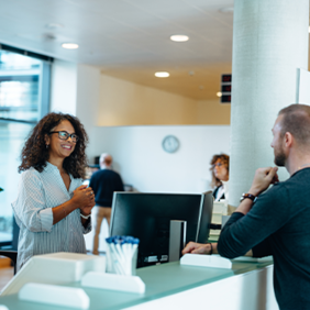 In einem modernen, hellen Bürogebäude stehen drei Personen an einem Empfangstresen. Eine Frau mit lockigem Haar und Brille steht hinter dem Tresen und lächelt freundlich. Vor dem Tresen stehen ein Mann und eine Frau, die beide ebenfalls lächeln. Im Hintergrund sind weitere Personen zu sehen, die sich in der offenen und gut beleuchteten Umgebung bewegen. Pflanzen und moderne Möbel tragen zur angenehmen Atmosphäre bei.