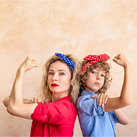 Zwei Frauen, die Rücken an Rücken stehen und ihre Arme in einer starken Pose beugen. Beide tragen bunte Haarbänder – eine Person trägt ein blaues Kopftuch mit weißen Punkten, die andere ein rotes Kopftuch mit weißen Punkten. Die Person links trägt ein rotes Hemd, während die Person rechts ein blaues Hemd trägt. Der Hintergrund ist neutral beige gehalten. Die beiden Personen schauen entschlossen in die Kamera.