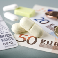 Auf dem Bild sind eine Krankenversicherungskarte, mehrere Euro-Banknoten und -Münzen sowie einige Tabletten zu sehen. Die Krankenversicherungskarte liegt links im Vordergrund, während die Banknoten und Münzen rechts daneben platziert sind. Die Tabletten befinden sich in der Mitte des Bildes.