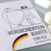Auf dem Bild sind eine Krankenversicherungskarte, mehrere Euro-Banknoten und -Münzen sowie einige Tabletten zu sehen. Die Krankenversicherungskarte liegt links im Vordergrund, während die Banknoten und Münzen rechts daneben platziert sind. Die Tabletten befinden sich in der Mitte des Bildes.