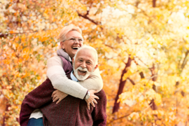 Ein älteres Paar genießt einen sonnigen Herbsttag im Freien. Die Frau, die eine Brille trägt, umarmt den Mann von hinten und beide lachen herzlich. Sie sind von herbstlich gefärbten Bäumen umgeben, deren Blätter in warmen Gelb- und Orangetönen leuchten. Der Mann trägt einen weinroten Pullover, während die Frau eine helle Jacke anhat. 