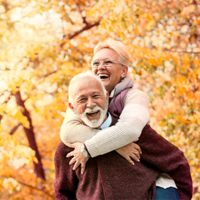 Ein älteres Paar genießt einen sonnigen Herbsttag im Freien. Die Frau, die eine Brille trägt, umarmt den Mann von hinten und beide lachen herzlich. Sie sind von herbstlich gefärbten Bäumen umgeben, deren Blätter in warmen Gelb- und Orangetönen leuchten. Der Mann trägt einen weinroten Pullover, während die Frau eine helle Jacke anhat. 