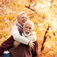 Ein älteres Paar genießt einen sonnigen Herbsttag im Freien. Die Frau, die eine Brille trägt, umarmt den Mann von hinten und beide lachen herzlich. Sie sind von herbstlich gefärbten Bäumen umgeben, deren Blätter in warmen Gelb- und Orangetönen leuchten. Der Mann trägt einen weinroten Pullover, während die Frau eine helle Jacke anhat. 