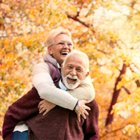 Ein älteres Paar genießt einen sonnigen Herbsttag im Freien. Die Frau, die eine Brille trägt, umarmt den Mann von hinten und beide lachen herzlich. Sie sind von herbstlich gefärbten Bäumen umgeben, deren Blätter in warmen Gelb- und Orangetönen leuchten. Der Mann trägt einen weinroten Pullover, während die Frau eine helle Jacke anhat. 